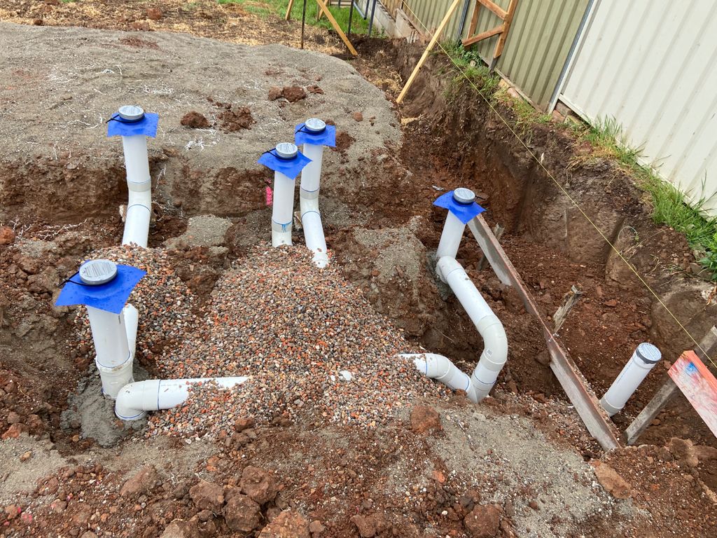 F7CF3D5F-363F-4A21-8EA3-3F267F8F7817 Trench with white PVC plumbing pipes capped with blue tape emerging from dirt, surrounded by earth and gravel, indicating construction work.